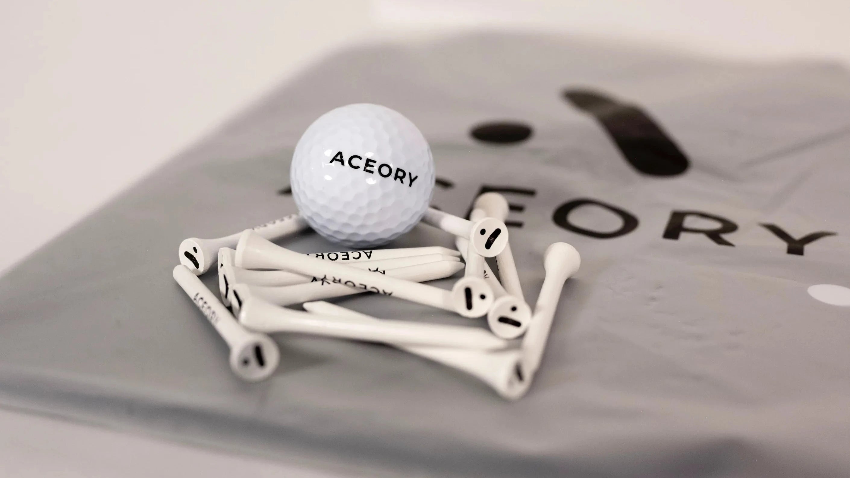Aceory branded golf ball and white tees placed on Aceory packaging bag, minimalist studio close-up.