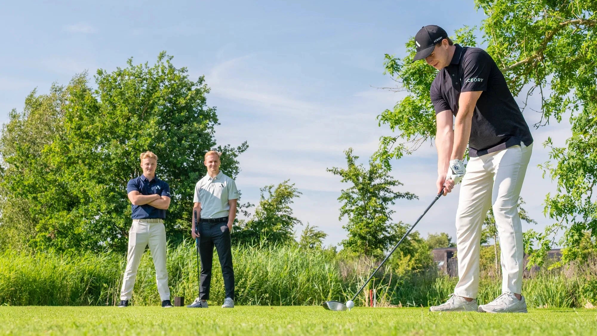 Golfer in Aceory polo teeing off while two teammates watch on a sunny fairway.