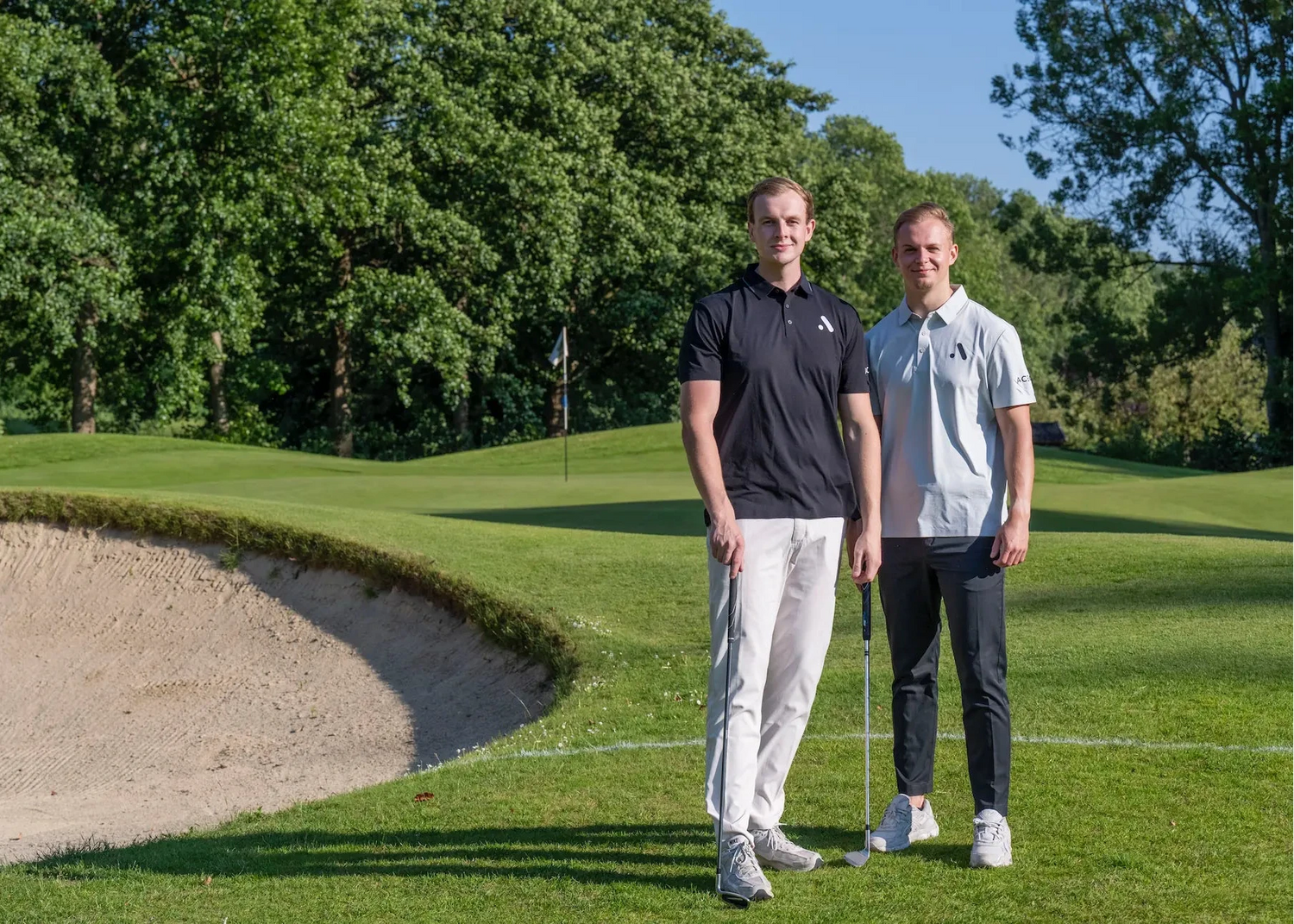 Two golfers standing beside a bunker wearing black and light grey Aceory polos under bright afternoon light.